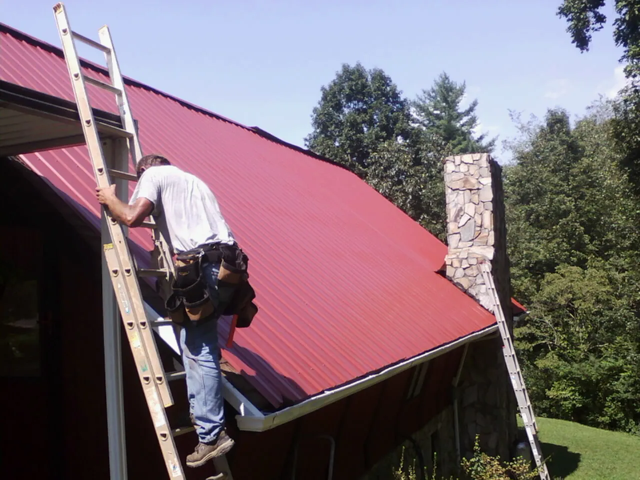 Roofer on a ladder beside a red metal roof and stone chimney — Soffit & Fascia Repair by Roofing Brothers in Faucett